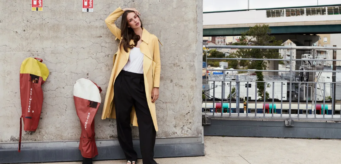 Trench jaune moutarde et pantalon noir, cheveux longs détachés, décor urbain industriel, collection PE 2026